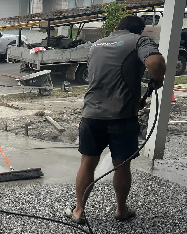 A DCF worker in a grey DCF Concreting shirt hosing down a new exposed aggregate driveway, with a tray truck and a ute parked behind
