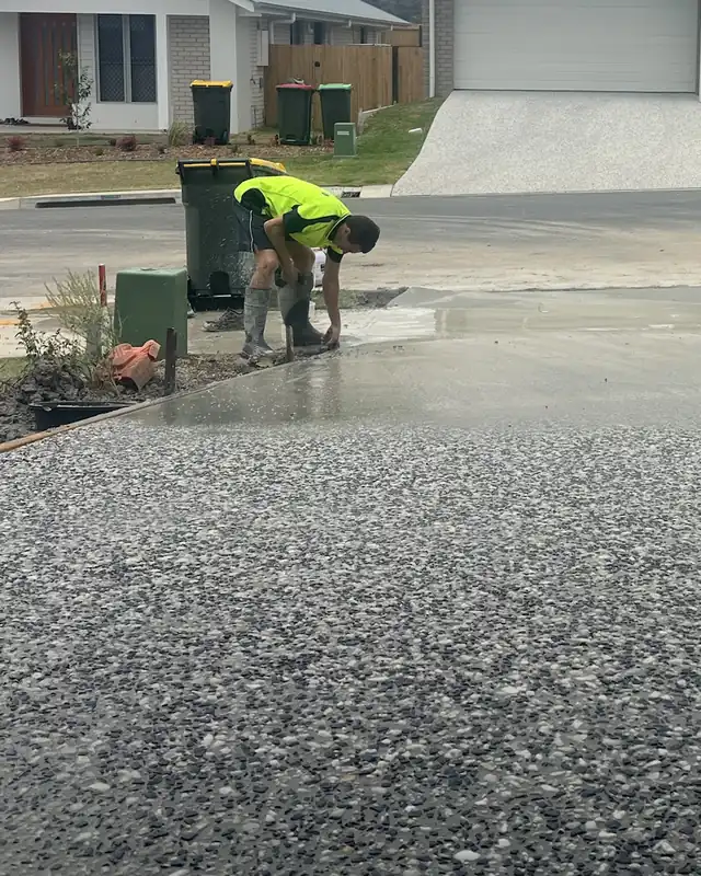 A worker in hi-vis bent over finishing the edge of a fresh salt and pepper exposed aggregate driveway, with new homes behind