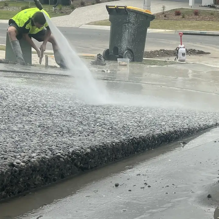 A jet of water washing the cement film off fresh exposed aggregate to bring up the stone, with a clean stepped edge down to a plain slab and a DCF worker in hi-vis on the kerb behind